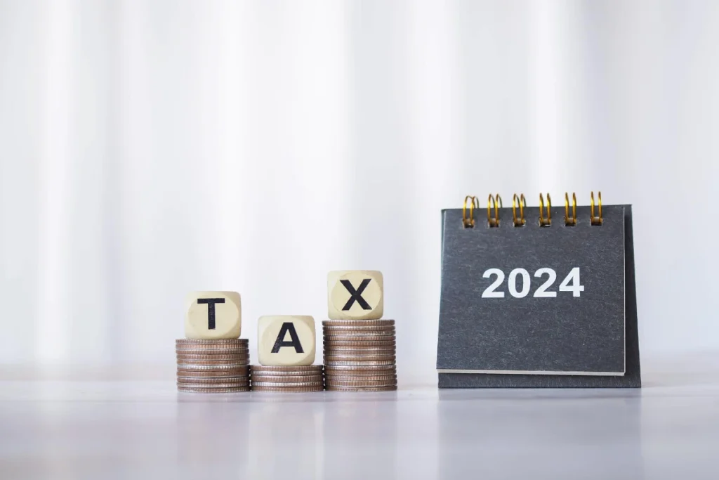 2024 desk calendar, wooden block with 'tax,' and coins stack symbolize saving, tax payment, financial investment, and business growth in 2024.